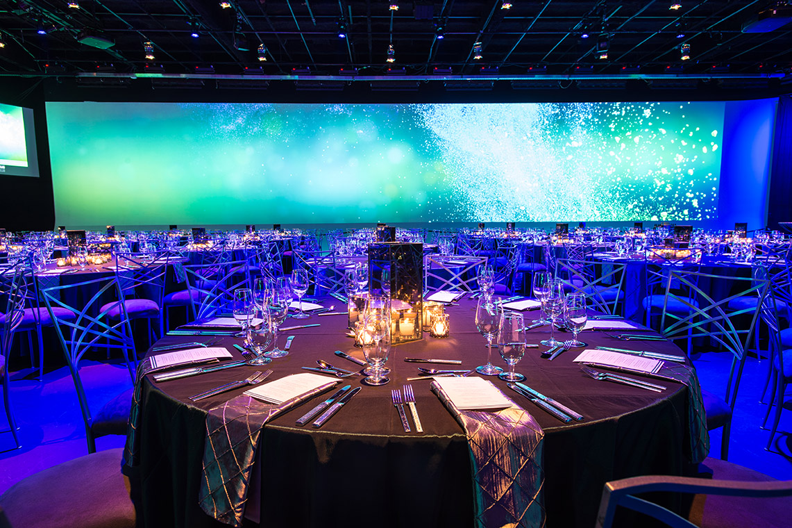 Large dimly lit conference room with circular dining tables and a bright digital screen on the rear wall.