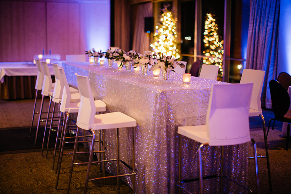 Rectangular table in a colorfully lit room with flowers and candles on top surrounded by white seats.
