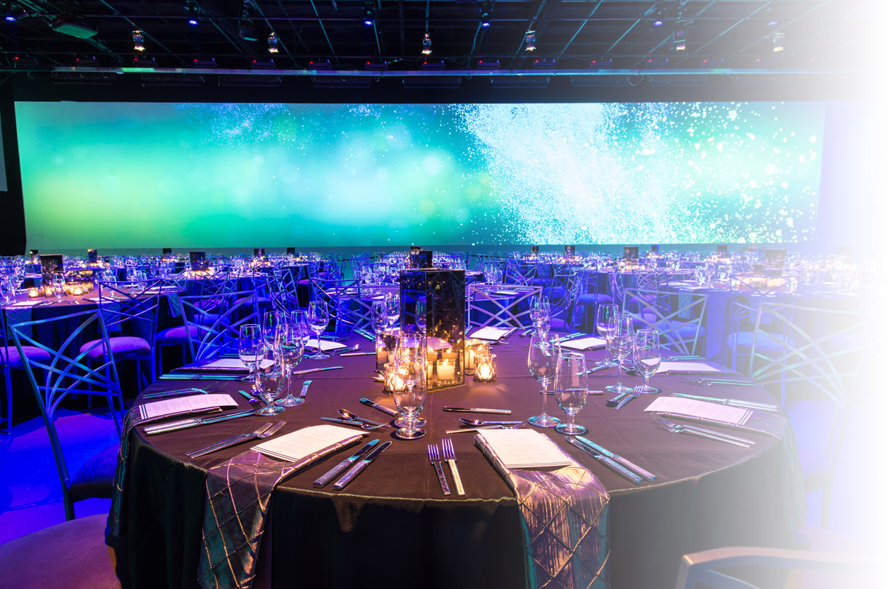 Large dimly lit conference room with circular dining tables and a bright digital screen on the rear wall.