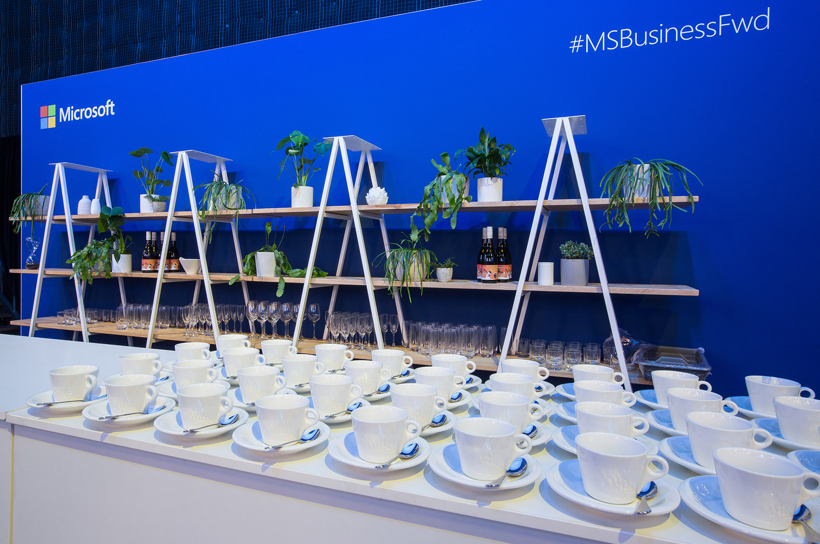 Table full of coffee cups and spoons in front of a blue Microsoft branded backdrop with greenery.
